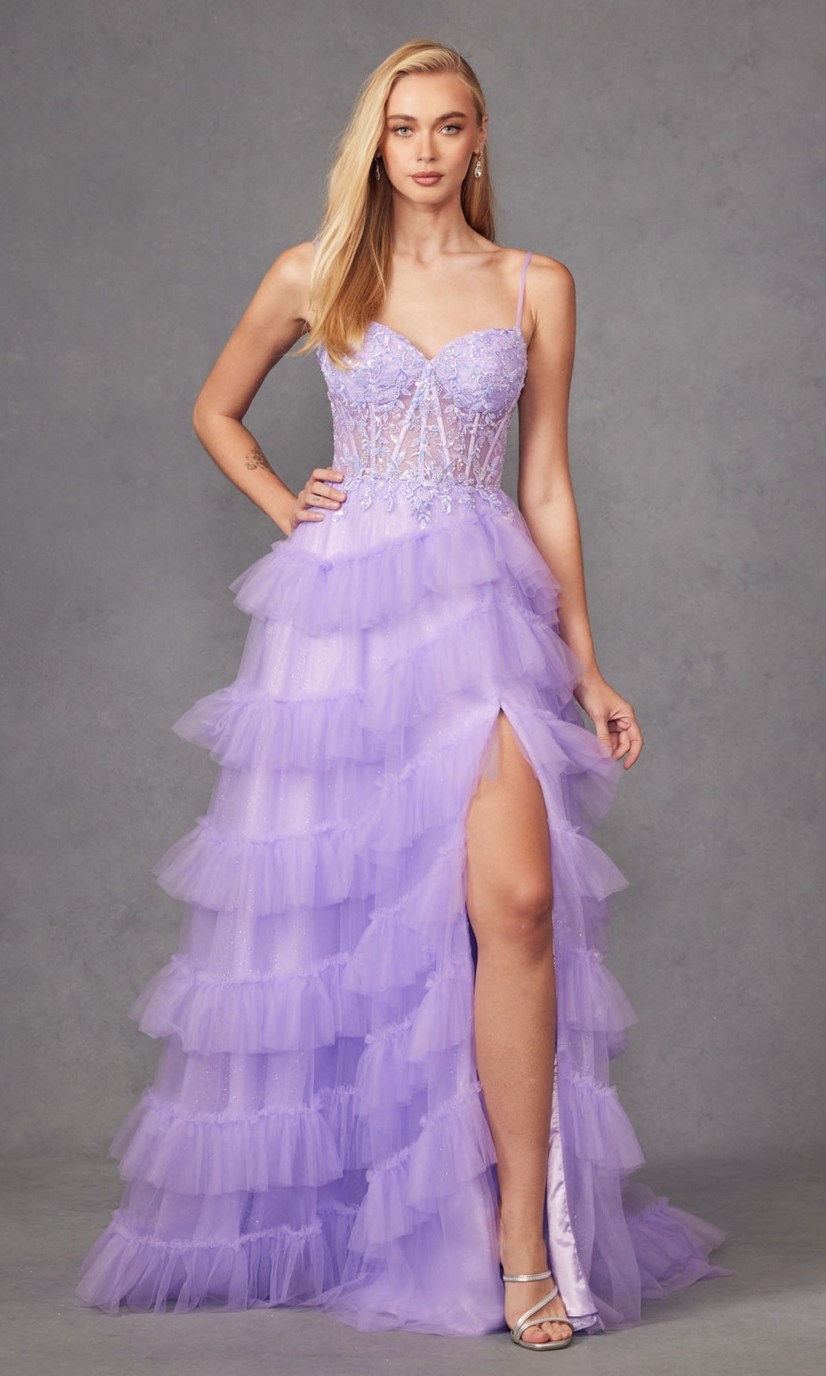 A woman stands against a gray background wearing the Long Prom Dress JT2459A by Juliet—a pastel purple ruffle tulle ball gown featuring a sheer beaded bodice, spaghetti straps, and a high slit.